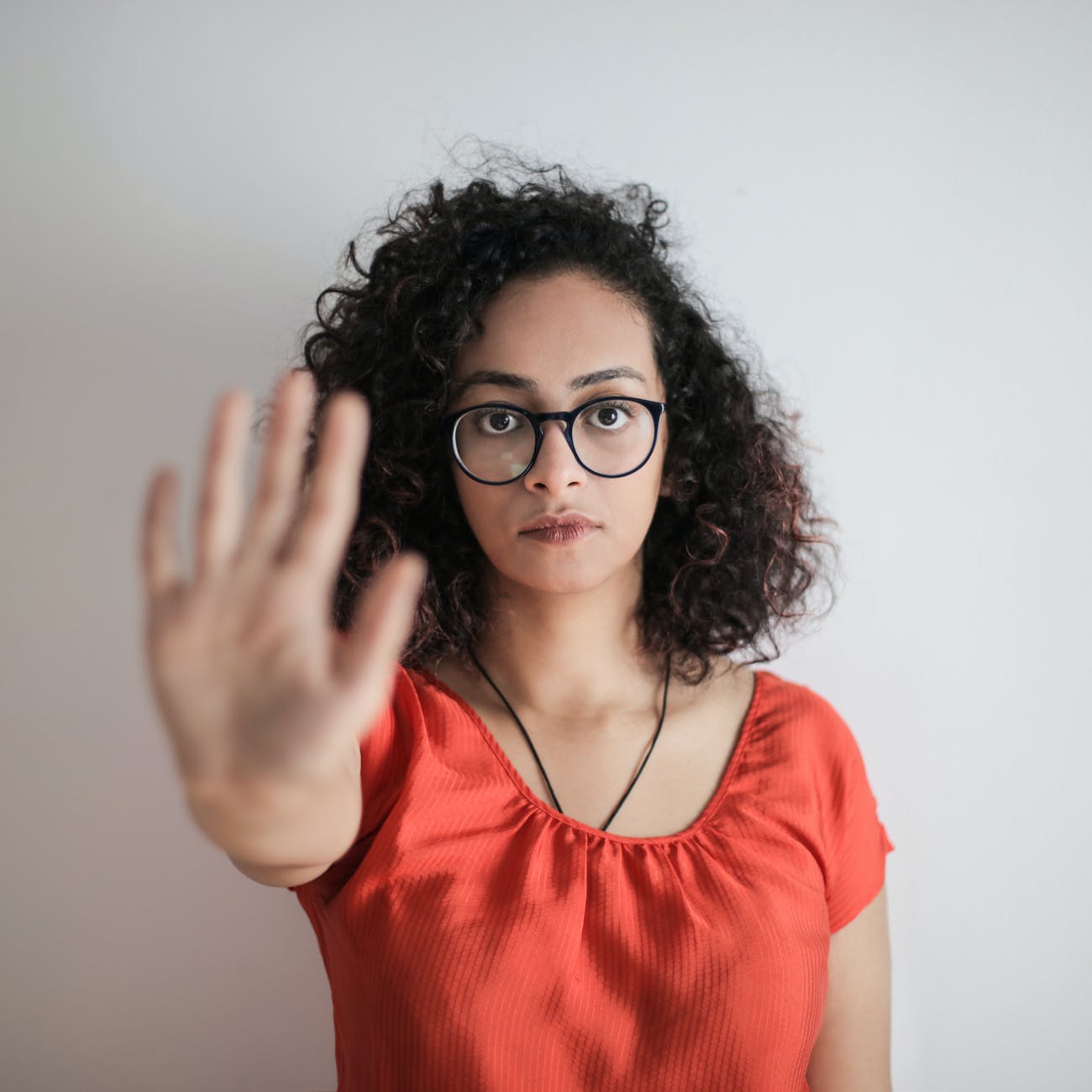 portrait photo of woman in red top wearing black framed eyeglasses holding out her hand in stop gesture